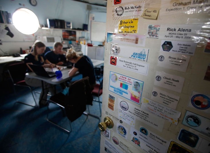 Photograph of an MDRS crew gathered around a table during a planning session, showing six crew members reviewing documents and maps